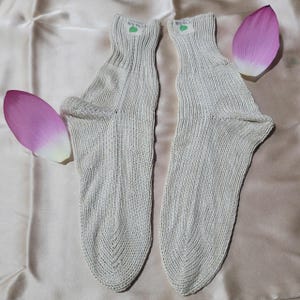 May include: Two light beige knitted socks with ribbed cuffs, each with a small label with green text. Pink lotus petals are placed near the socks on a satin surface. The socks appear to be made of a soft, natural fibre.