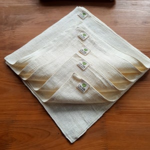 May include: A stack of five off-white, square cloth napkins arranged in a diamond shape. Each napkin has a small label with the text "Chu Silk" and a leaf logo. The napkins are made of a textured fabric.