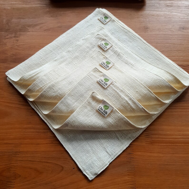 May include: A stack of five off-white, square cloth napkins arranged in a diamond shape. Each napkin has a small label with the text "Chu Silk" and a leaf logo. The napkins are made of a textured fabric.