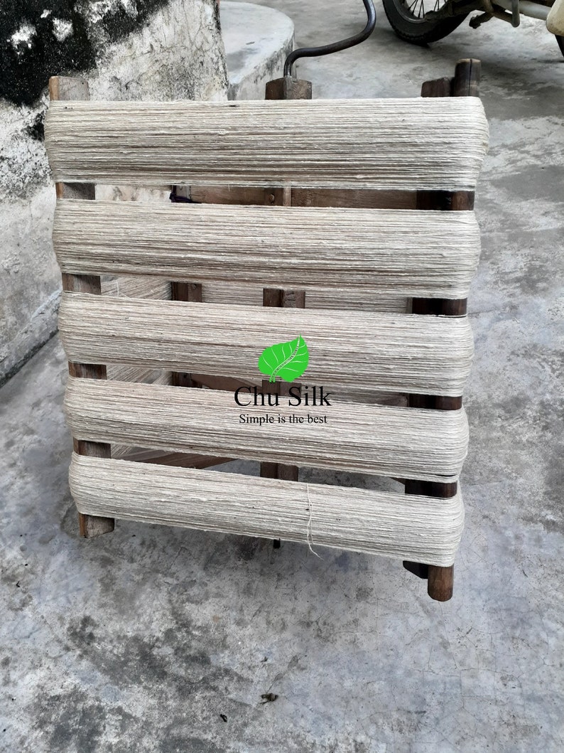 May include: A wooden yarn swift with tightly wound, off-white yarn. The swift has a rectangular frame with horizontal wooden bars and vertical supports. The text "Chu Silk Simple is the best" is visible on the bottom right.