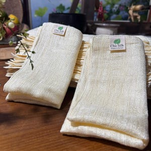 May include: Two folded, cream-colored silk cloths with a textured weave are displayed. Each cloth has a small label that reads "Chu Silk" with a leaf graphic and the text "Simple is the best". The cloths are stacked on a wooden surface.