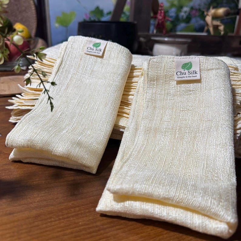 May include: Two folded, cream-colored silk cloths with a textured weave are displayed. Each cloth has a small label that reads "Chu Silk" with a leaf graphic and the text "Simple is the best". The cloths are stacked on a wooden surface.
