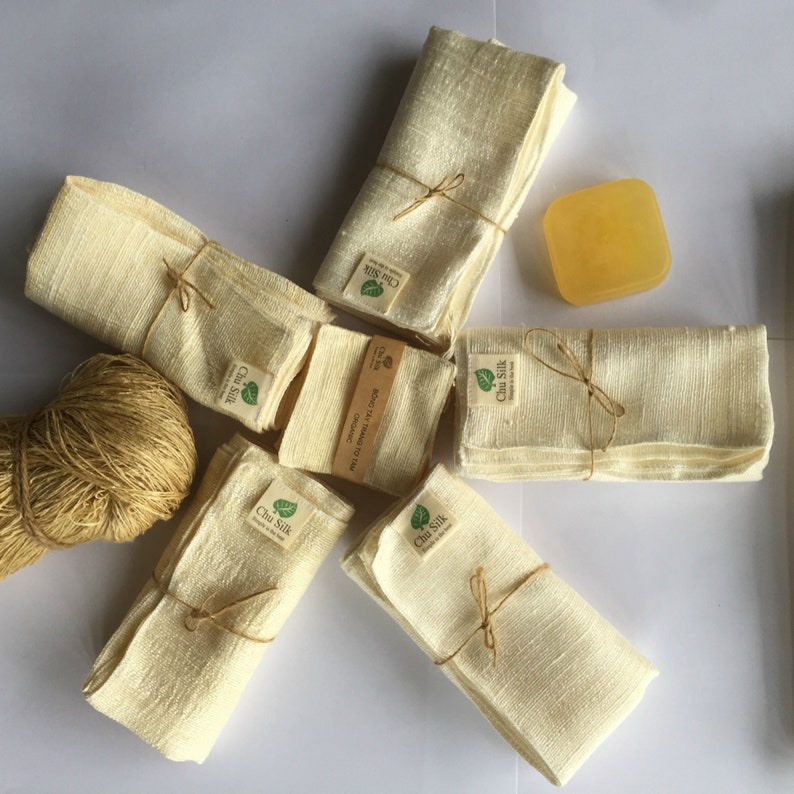 May include: Arrangement of cream-colored silk cloths, some folded and tied with twine, and a square yellow soap. Each cloth has a label that reads "Chu Silk". A ball of twine is also visible. The items are arranged on a white surface.