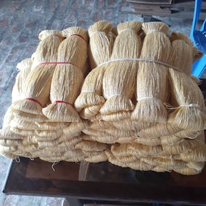 May include: A stack of bundles of golden-colored fiber, tightly bound with red and white bands. The bundles are stacked on a dark wooden table, suggesting a craft or textile material.