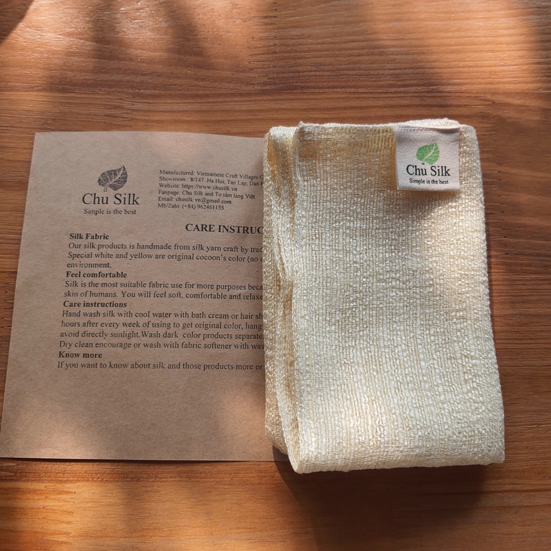 May include: A folded, cream-colored silk fabric with a textured surface, next to a brown paper card. The card features the text "Chu Silk" and care instructions. The fabric is made from silk yarn and is soft and comfortable.