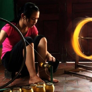 May include: A woman in a pink shirt and black pants is working with a spinning wheel. The image shows a close-up of the process of creating thread or yarn, with a blurred, glowing circle of material.