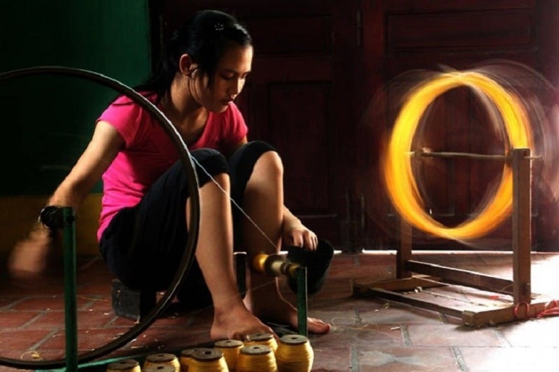 May include: A woman in a pink shirt and black pants is working with a spinning wheel. The image shows a close-up of the process of creating thread or yarn, with a blurred, glowing circle of material.