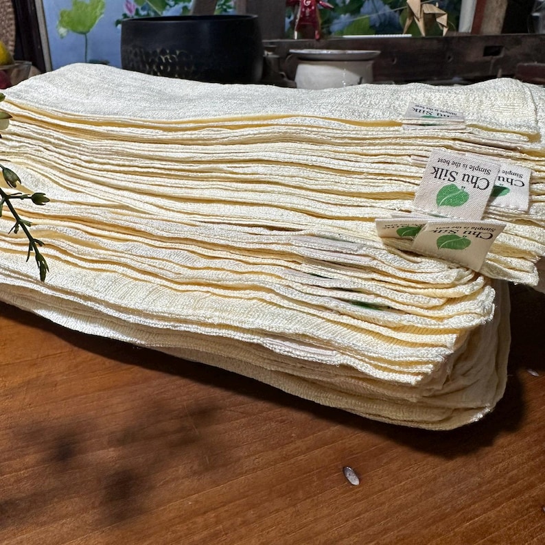 May include: A stack of folded, light yellow fabric cloths. The cloths have a textured appearance and are neatly stacked. Small tags with the text "Chu Silk" and a leaf logo are visible on some of the cloths. The cloths appear to be made of a natural fiber.