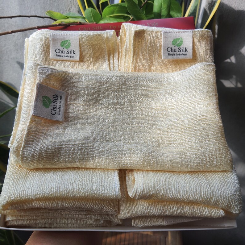 May include: A stack of folded, cream-colored silk cloths. Each cloth has a textured surface and a small label with the text "Chu Silk Simple is the best" and a green leaf logo. The cloths are neatly arranged in a white box.