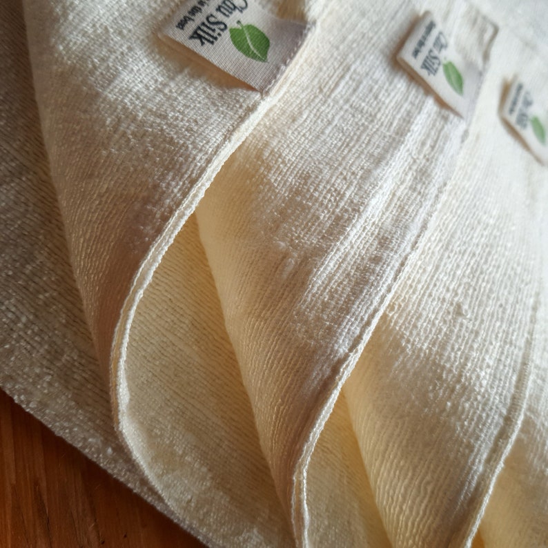 May include: Close-up of folded, off-white fabric with a textured weave. The fabric is layered, revealing the edges and seams. Small tags with the text "Chu Silk" and a green leaf are visible.