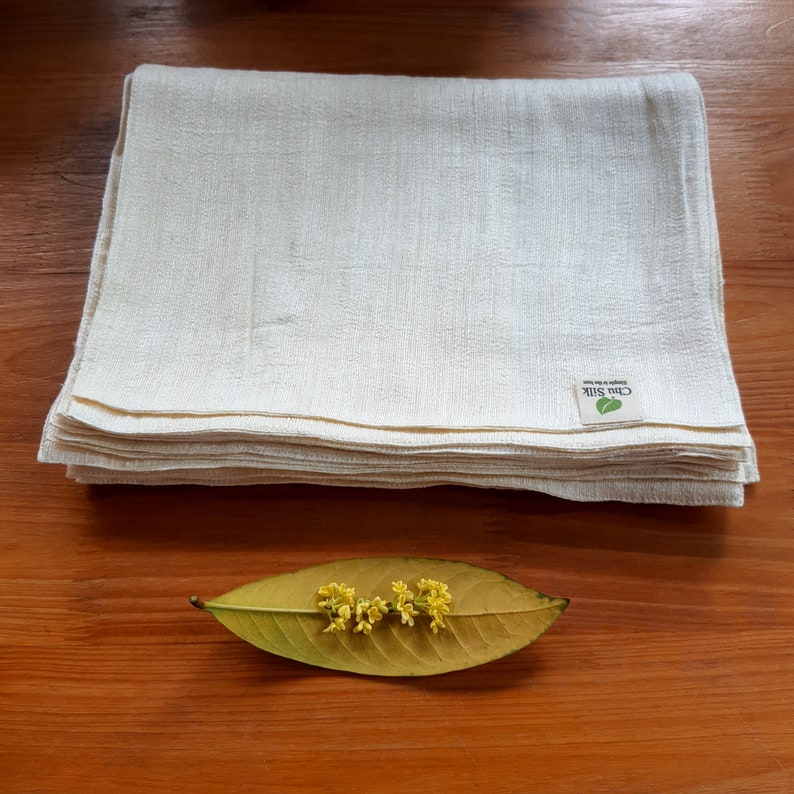 May include: A stack of off-white silk cloths with a textured weave. A small tag with the text "Chu Silk" and a green leaf logo is visible. A yellow leaf with small yellow flowers rests on a wooden surface.