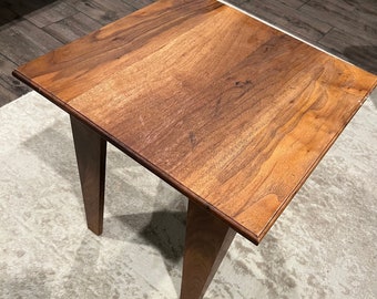 May include: A small, square wooden end table with a light brown wood finish. The table has four legs that are angled inwards.