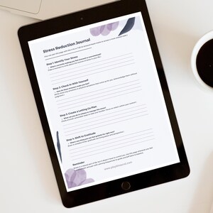 Op de afbeelding: Een digitale tablet met een afdrukbaar stressreductie-dagboek met stappen om stress te identificeren, bij jezelf na te gaan, een loslaatplan te maken en je te richten op dankbaarheid. Het dagboek ligt op een wit oppervlak met een kopje koffie en een witte pen.