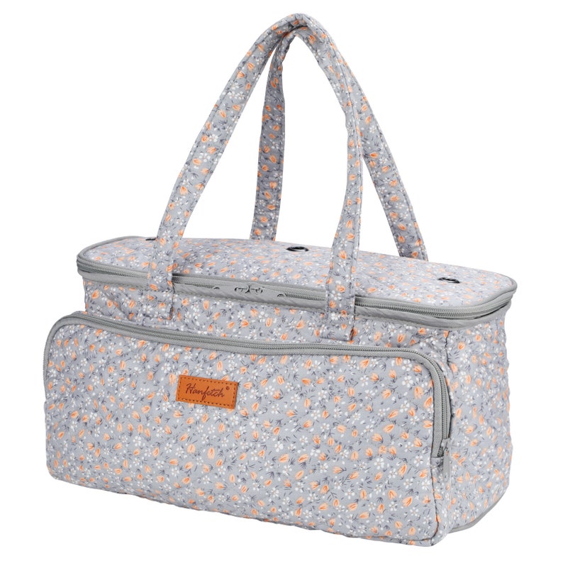 May include: A gray quilted tote bag with a floral print featuring small orange flowers. The bag has two top handles and a zipper closure. The bag has a brown leather patch with the text "Handstitch" on it.