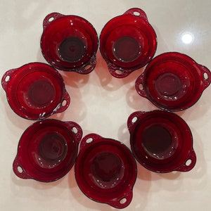 Puede incluir: Un conjunto de siete cuencos de vidrio rojo rubí dispuestos en un patrón circular. Cada cuenco tiene dos asas y un borde decorativo. Los cuencos están hechos de vidrio rojo transparente.