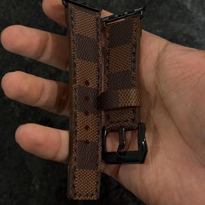 May include: Brown and tan checkered Apple Watch band with a black buckle. The band is held in a hand, showcasing the pattern and design. The band is made of a durable material.