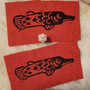 May include: Two rectangular red fabric patches featuring a black block-print style illustration of a fish. A white twenty-sided die with black numbers is positioned between the two patches for scale, showing the number 20 on its top face.