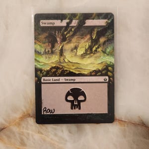Puede incluir: Una carta coleccionable de Magic: The Gathering titulada Swamp, con una ilustración de paisaje oscuro y atmosférico con árboles retorcidos y niebla verde. La carta incluye el texto Swamp, Basic Land - Swamp, un símbolo de calavera negra y la firma Row.