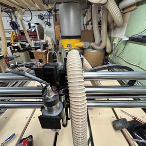 以下が含まれることがあります： ダストコレクションシステムを搭載したCNCマシンのクローズアップ。マシンには、黒と銀のスピンドルに接続された白と灰色のホースが付いています。マシンは薄い茶色の表面の上に置かれています。