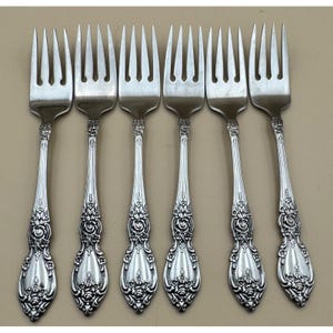 May include: Six silver forks with ornate floral and scrollwork detailing on the handles. The tines are evenly spaced. The forks are arranged in a row, showcasing their elegant design, perfect for formal dining.