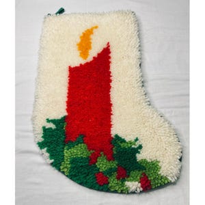 May include: A cream-colored latch hook Christmas stocking featuring a central red candle with a yellow flame. The base of the stocking is decorated with green holly leaves and small red berries, all rendered in a textured, looped yarn pile.