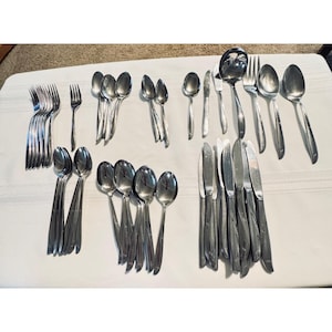 May include: A large collection of stainless steel flatware including various forks, spoons, knives, and a soup ladle. The set features a polished metallic finish with simple, smooth handles arranged in groups across the surface.