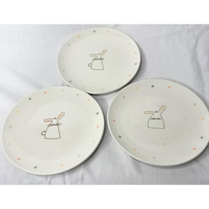 May include: Three white ceramic plates with a whimsical bunny design. Each plate features a hand-drawn bunny illustration in the center and is encircled by small, colorful polka dots. The plates are suitable for serving food.