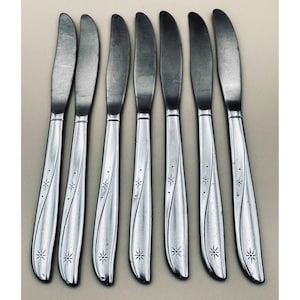 May include: Set of eight stainless steel butter knives. Each knife has a curved blade and a handle with a decorative starburst pattern. The knives are arranged in a row, with the blades facing up.