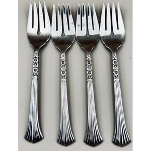 May include: Four silver forks with ornate floral and bow designs on the handles. The tines are long and slender. The handles have a fluted design that flares out at the base. The forks are arranged in a row against a neutral background.