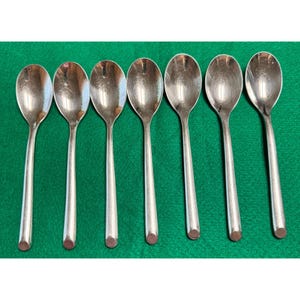 May include: Seven silver spoons with oval bowls and long, slender handles are arranged on a green surface. The spoons are evenly spaced, reflecting light. This set of spoons is suitable for serving.