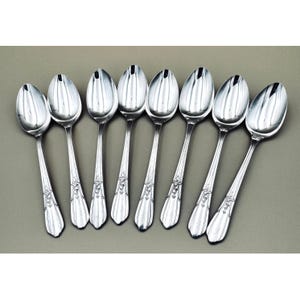 May include: A set of eight silver spoons arranged on a gray surface. The spoons have an oval bowl shape with a central ridge and a decorative design on the handle. The spoons are likely for serving or eating.