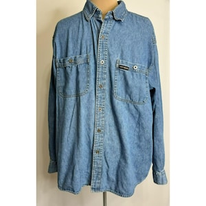 May include: Light blue denim shirt with two chest pockets and a small logo patch. The long-sleeved shirt has a button-down front, a collar, and buttoned cuffs. The shirt is made of a medium-weight denim fabric.