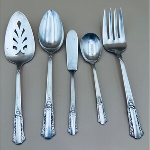 May include: A set of vintage silver-colored flatware, including a slotted serving spoon, a large spoon, a butter knife, a smaller spoon, and a fork. Each piece has a decorative handle with a floral design.