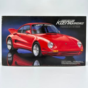 以下が含まれることがあります： 赤いケーニッヒ・スペシャルズ ポルシェ 911 ビターボのモデルカーキット。箱のイラストは、黒いフロントガラスとフロントガラスに黒いケーニッヒ・スペシャルズの文字が書かれた赤い車です。箱には、「Maquette a assembler」と「Unassembled Model Kit」の文字も書かれています。
