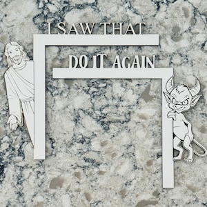Puede incluir: Un letrero de madera blanco con el texto "I SAW THAT DO IT AGAIN". El letrero presenta una representación de dibujos animados de Jesús a la izquierda y un diablo a la derecha, asomándose por los bordes del letrero.