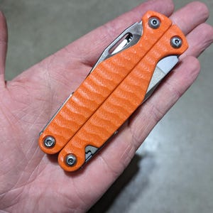 Puede incluir: Una pequeña multiherramienta naranja con un mango texturizado y partes metálicas plateadas. La herramienta está cerrada y se ve una pequeña cuchilla.