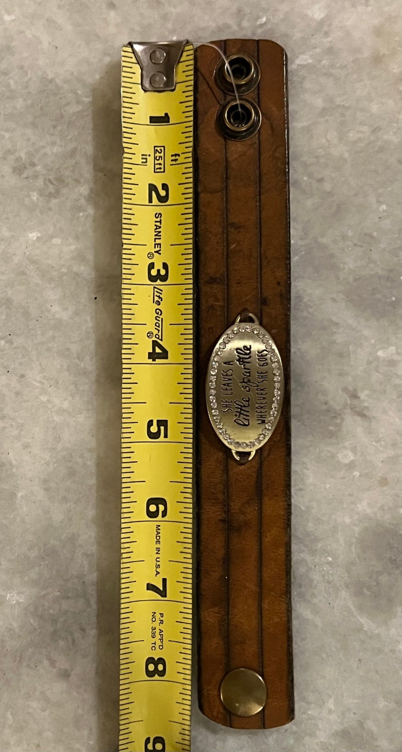 May include: A brown leather bracelet with a silver charm that reads "She leaves a little sparkle wherever she goes". The charm is decorated with small rhinestones. The bracelet is 7 inches long.