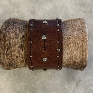 May include: A brown leather bracelet with silver studs and clear rhinestones. The bracelet is wrapped in a brown and white cowhide.