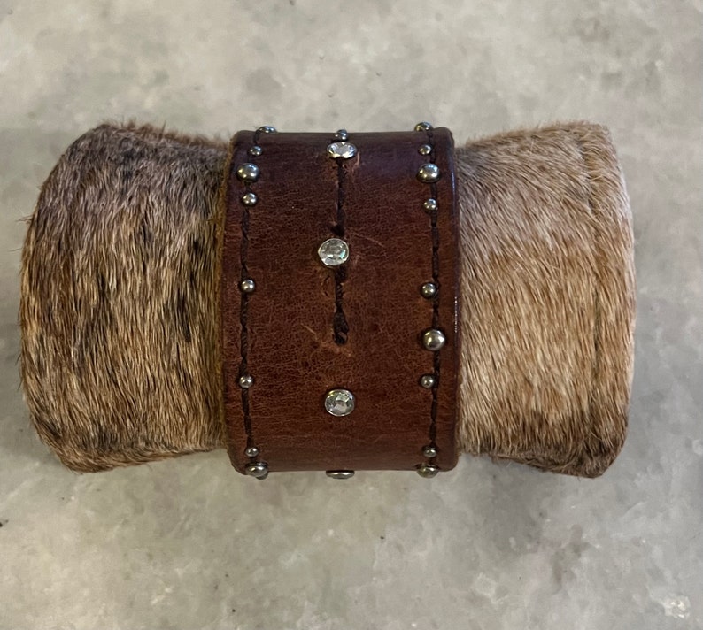 May include: A brown leather bracelet with silver studs and clear rhinestones. The bracelet is wrapped in a brown and white cowhide.