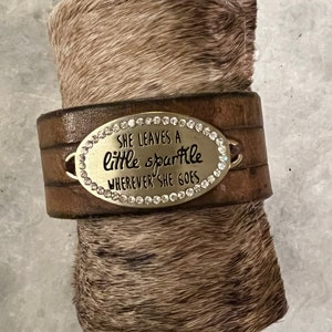 May include: Brown leather bracelet with a silver oval charm that says "She leaves a little sparkle wherever she goes". The charm is decorated with rhinestones.