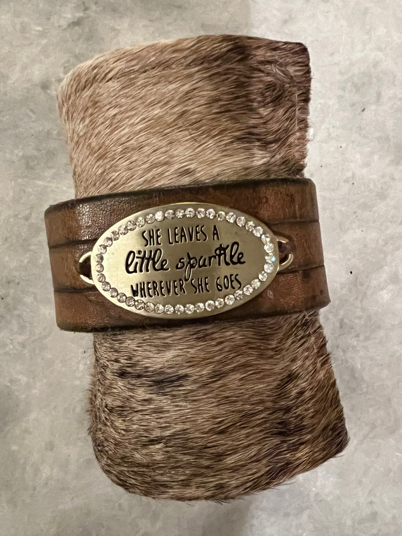May include: Brown leather bracelet with a silver oval charm that says "She leaves a little sparkle wherever she goes". The charm is decorated with rhinestones.