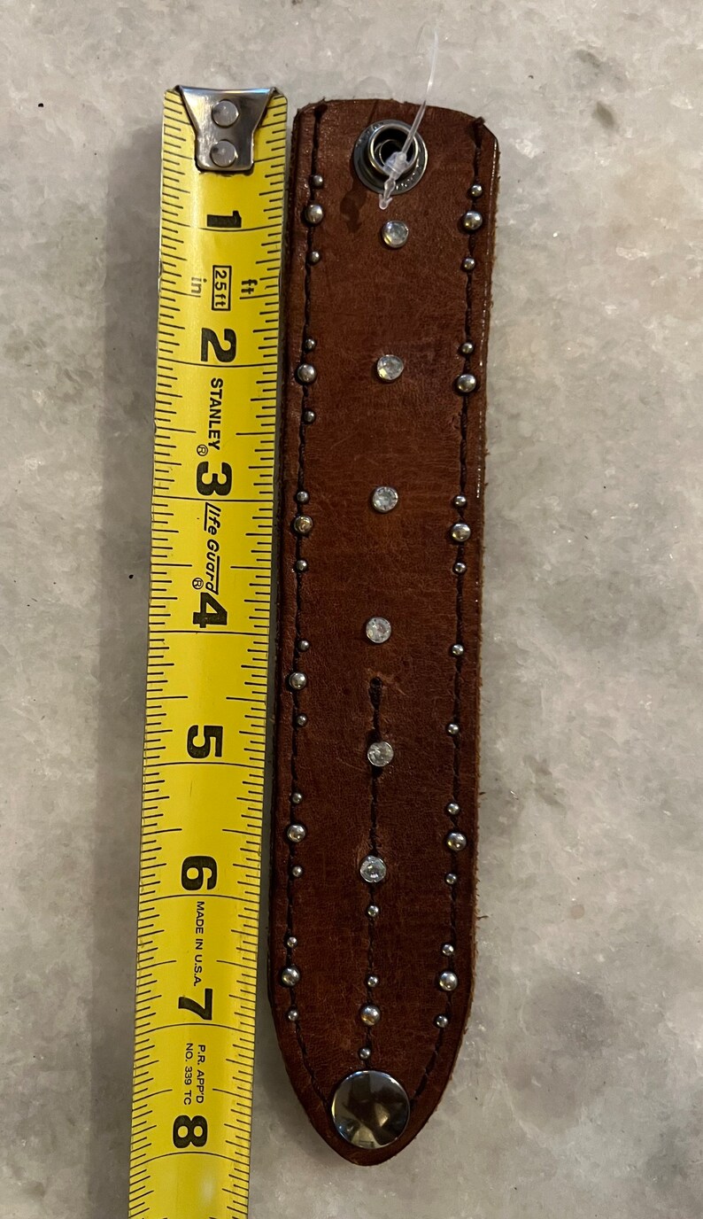 May include: Brown leather bracelet with silver studs and rhinestones. The bracelet is approximately 7 inches long.
