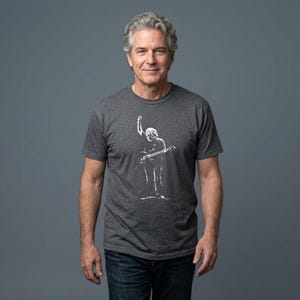 May include: A charcoal gray t-shirt featuring a white graphic of a musician playing a guitar with their fist raised. The t-shirt is worn by a person with gray hair and blue jeans.