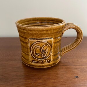 May include: A brown ceramic mug with a handle. The mug has a textured surface and a square emblem with the letters "CG" and the text "WACO TX". The mug is designed for hot beverages.