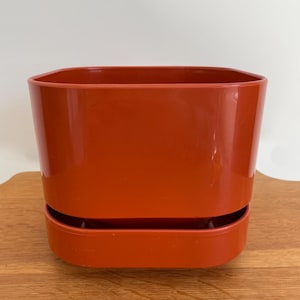 May include: A terracotta-colored plastic plant pot with a square top and a matching saucer base. The pot has a glossy finish and is sitting on a wooden surface.