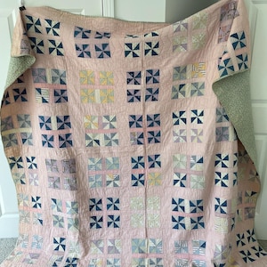 May include: A large rectangular quilt featuring a repeating pinwheel block pattern. The quilt is primarily light pink with individual blocks containing various blue, yellow, and white checkered or striped fabric squares. The reverse side is a light green fabric.