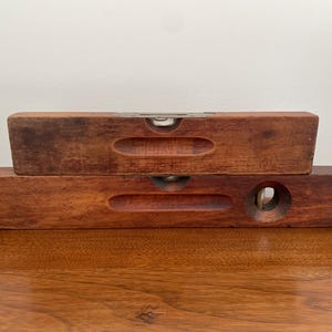 May include: Two vintage wooden levels, one stacked on top of the other. The levels are made of dark brown wood and have a rectangular shape with a curved indentation. Each level has a bubble vial for measuring levelness.