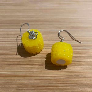 May include: A pair of dangle earrings featuring bright yellow corn cob charms. Each charm is textured to resemble individual kernels and is attached to a silver-toned metal hook. The earrings have a glossy finish and a small white circular base.