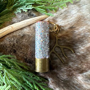 May include: A silver glitter-filled bullet casing keychain with a gold antler charm. The keychain is on a brown faux fur background.