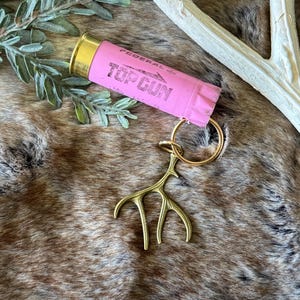 May include: A pink shotgun shell keychain with the text "TOP GUN" printed on it. The keychain has a gold-colored antler charm and a gold keyring. The background is a brown and tan textured surface.